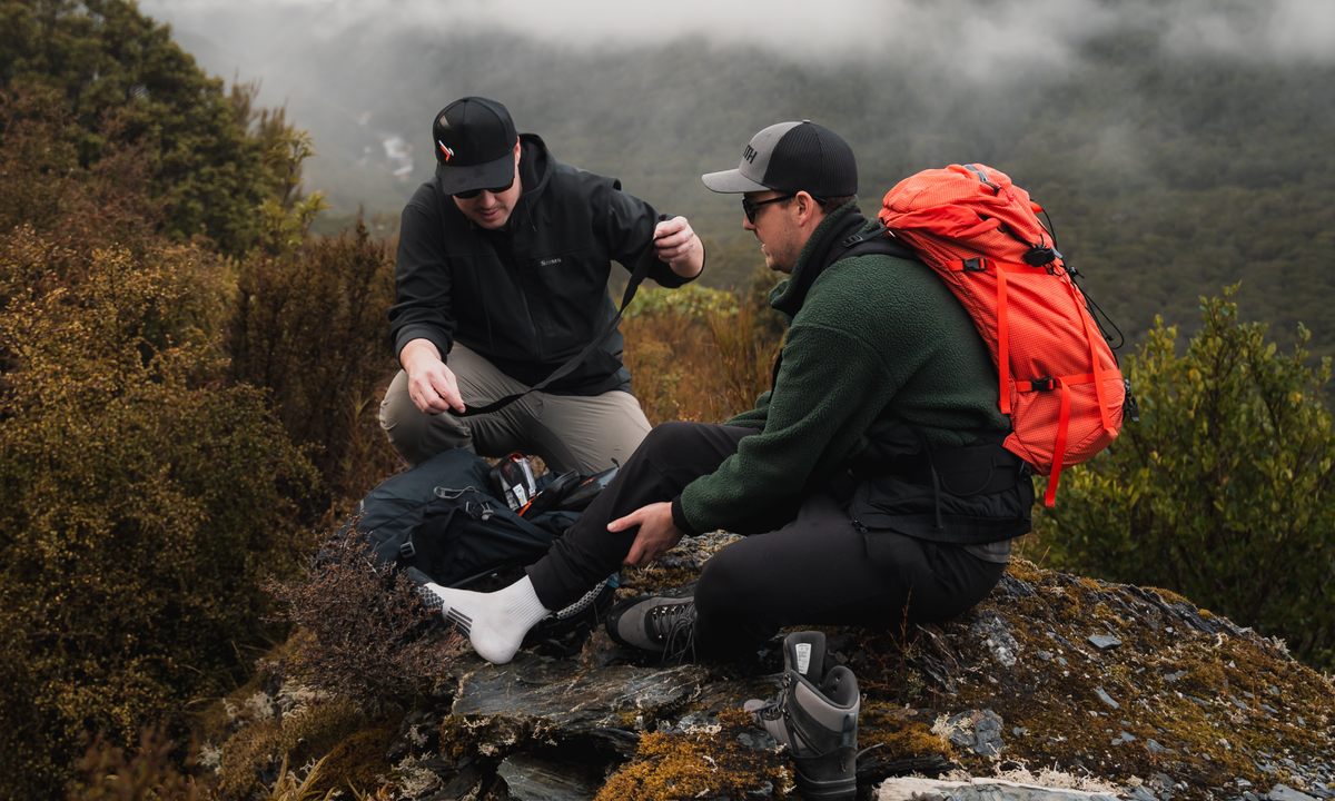 Two hikers checking an ankle on a New Zealand alpine trail, with an open KEA KIT beside them